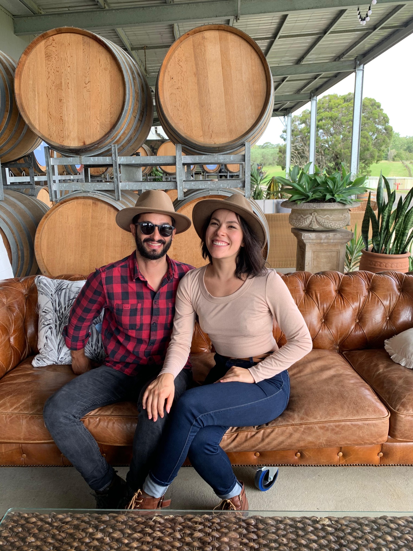 Carolina and Nacho smiling together on a leather couch at a winery, with wine barrels behind them